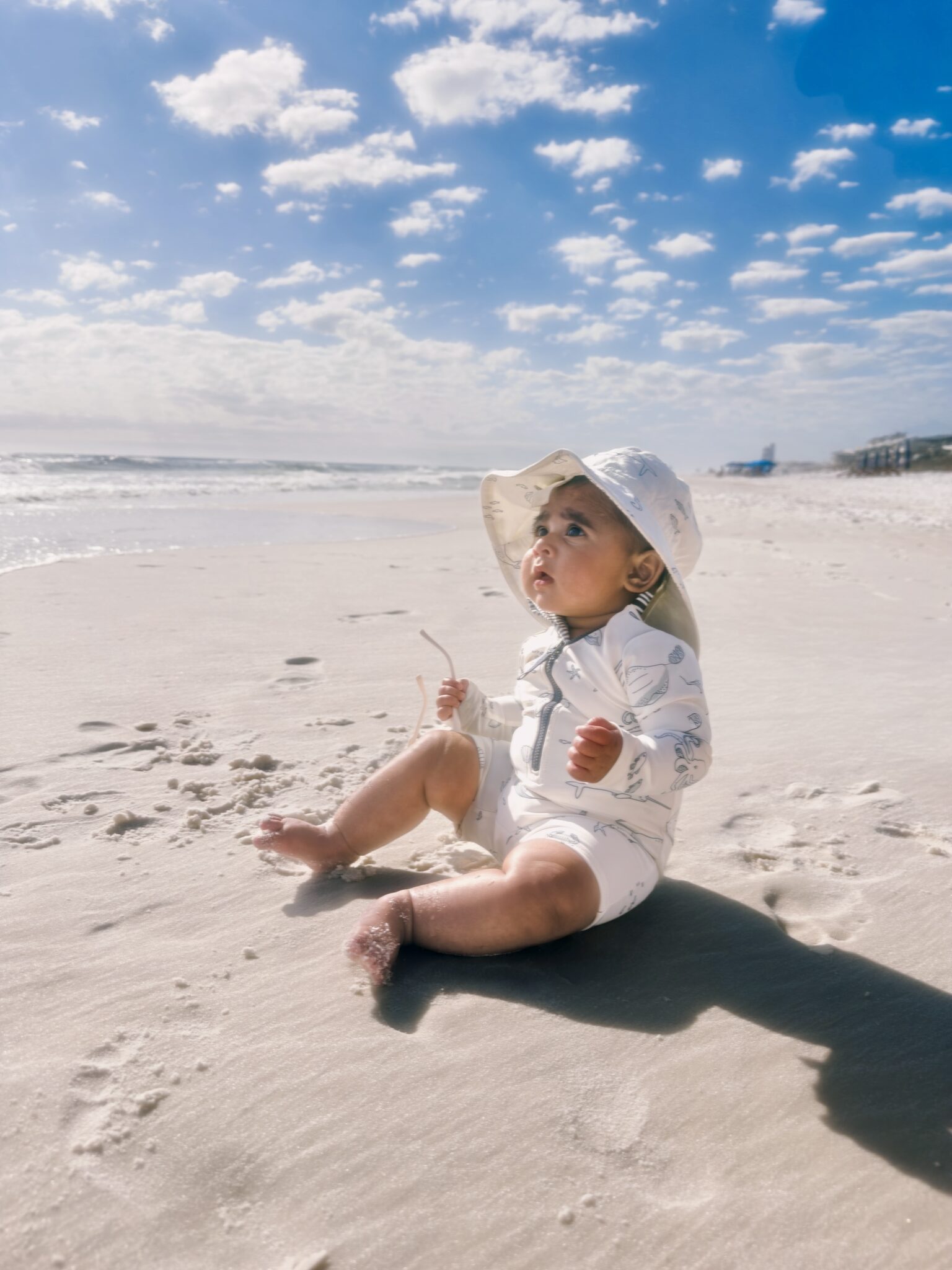 Arya in a sun hat sitting on white sand, meeting the ocean