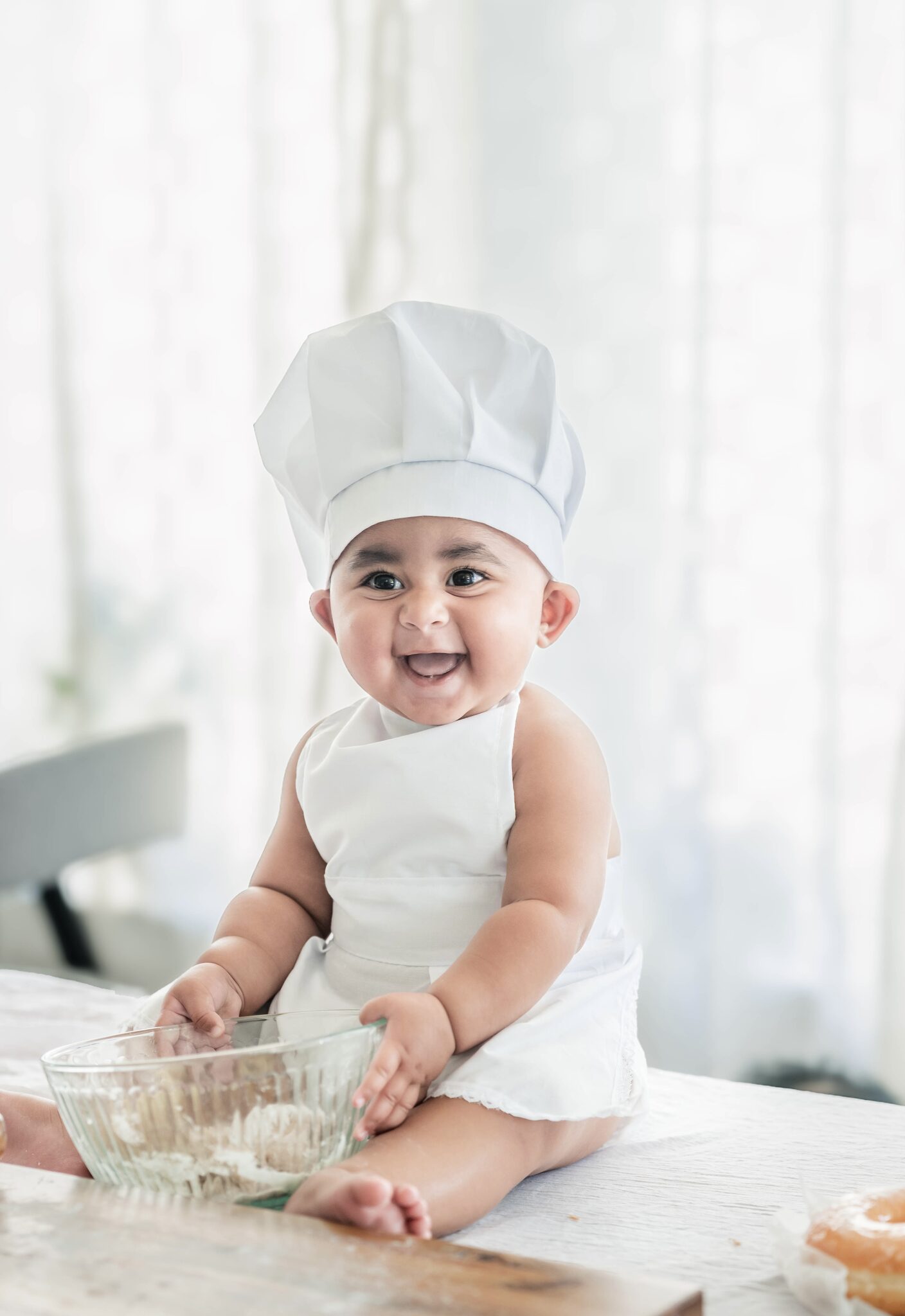 Arya in a tiny chef hat and apron, laughing with a mixing bowl